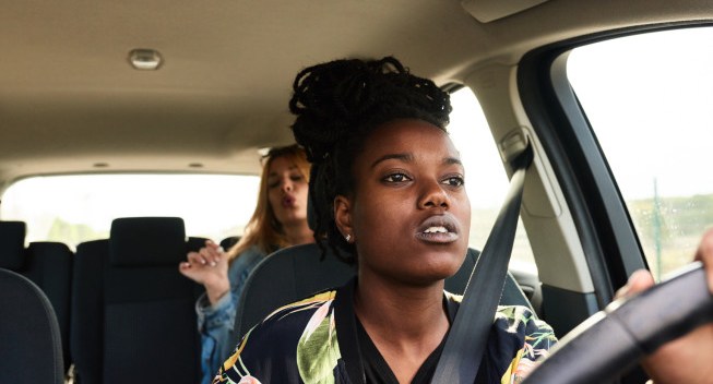 Portrait of concentrated girl driving car while her friend singing and having fun on backseat.