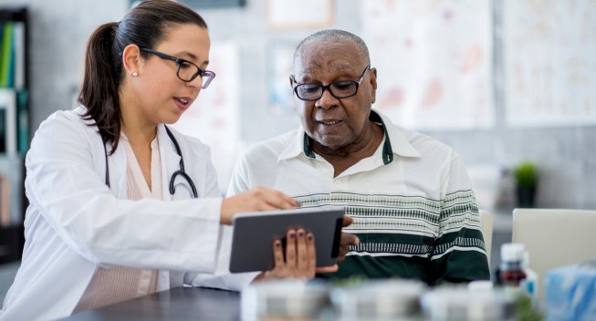 A senior man of African descent is indoors in a hospital room. He is watching his female doctor using a tablet computer. She is explaining a medication schedule to him.