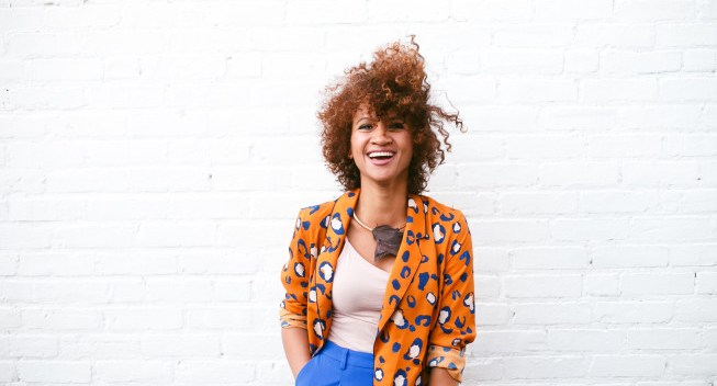 A young, beautiful woman with curly hair laughing against a white brick wall