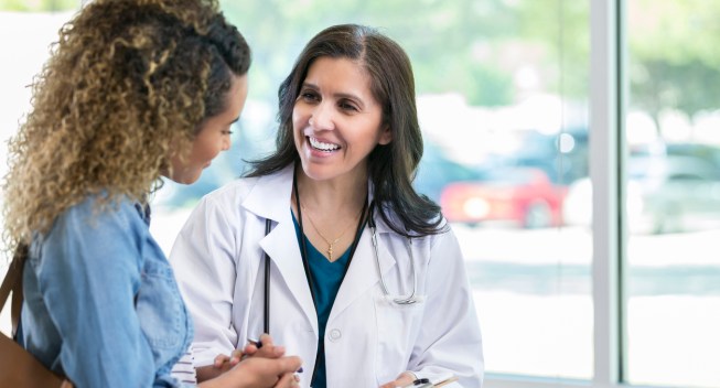 Caring mature female doctor shows test results to a young adult female patient. The doctor is smiling while talking with the patient.
