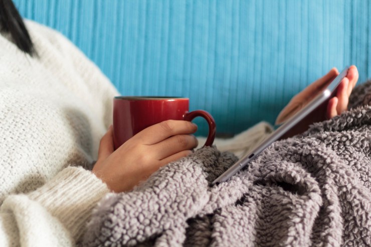 Woman bundled up in sweater and blanket drinking out of a mug while reading.