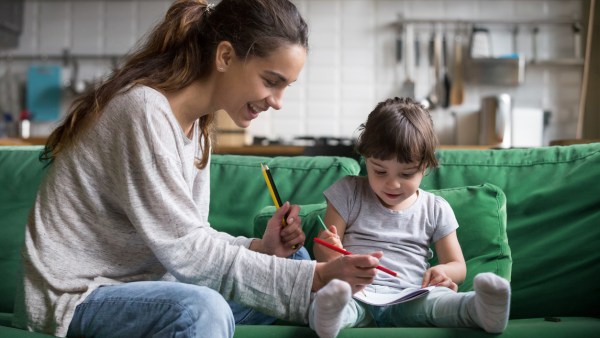 Smiling nanny and preschool kid girl drawing with colored pencils sitting on sofa together