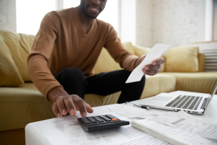 Young Black man using a calculator with paperwork and a laptop spread out in front of him.