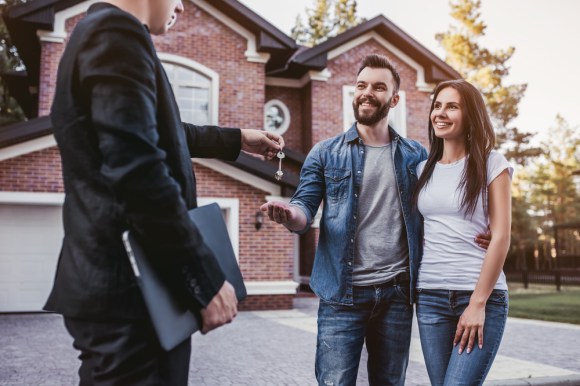 Realtor handing a young couple keys in front of a brick house.