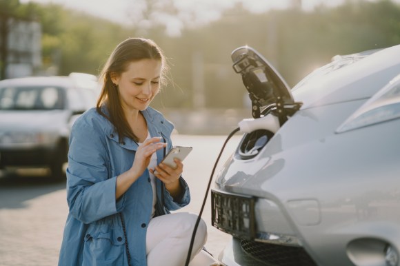Caucasian woman using her phone while charging her electric vehicle.