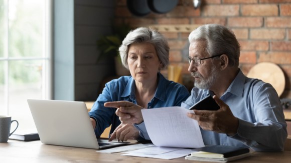 A focused senior couple sits at the table, looking at a laptop screen to pay bills and taxes online.