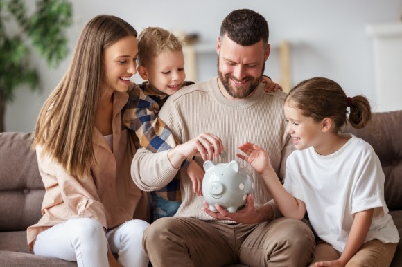 Cheerful parents with their kids smiling and putting coins into a piggy bank while sitting on the sofa.