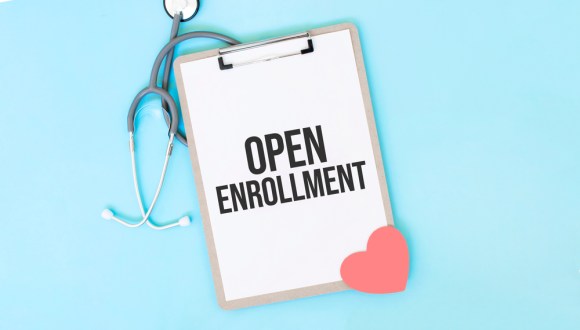 A clipboard with a paper that says “Open Enrollment” and a red heart rests on a stethoscope against a light blue background.