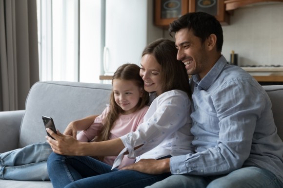 A couple and their daughter sit on the sofa using a smartphone to enjoy a video call with relatives.