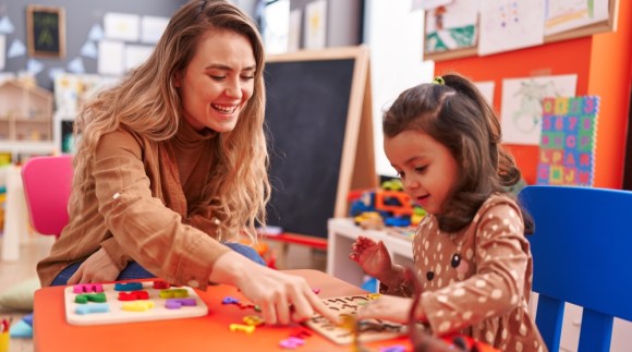 Young blonde woman doing a puzzle with a little girl in a daycare setting.