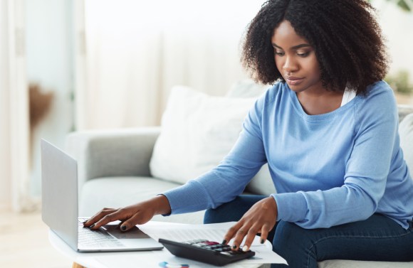 Black woman sitting on her couch using a calculator and typing on her laptop.