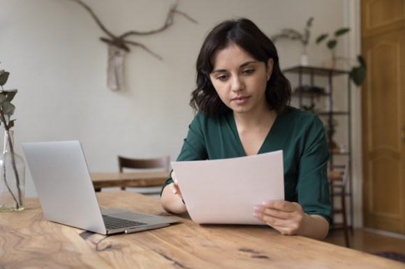  Young brunette woman reading a document with a laptop open in front of her.