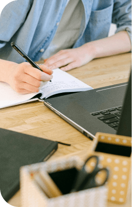 Photo of a person writing in a notebook at a desk in front of a laptop