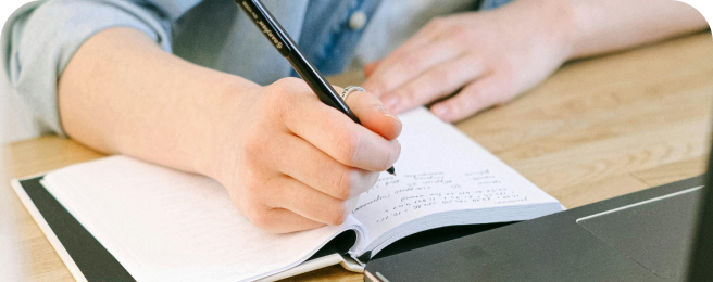 Photo of a person writing in a notebook at a desk in front of a laptop